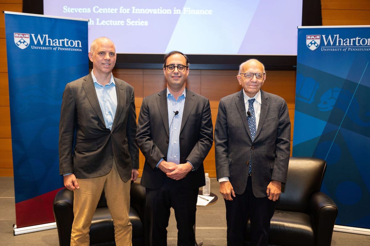 Professor David Musto, Professor Jeremy Siegel, and Matt Levine stand between two blue Wharton banners, smiling and posing toward the viewer