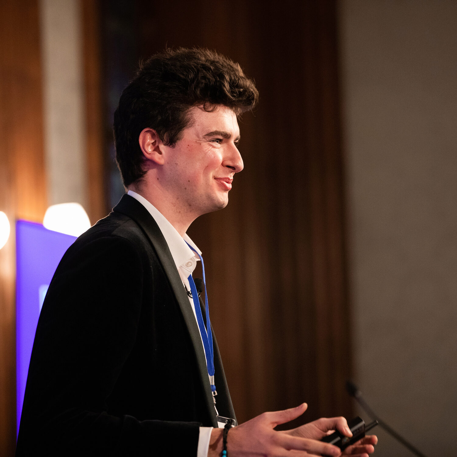 A person is giving a presentation, holding a remote and smiling. They are wearing formal attire with a lanyard, standing against a blurred background.