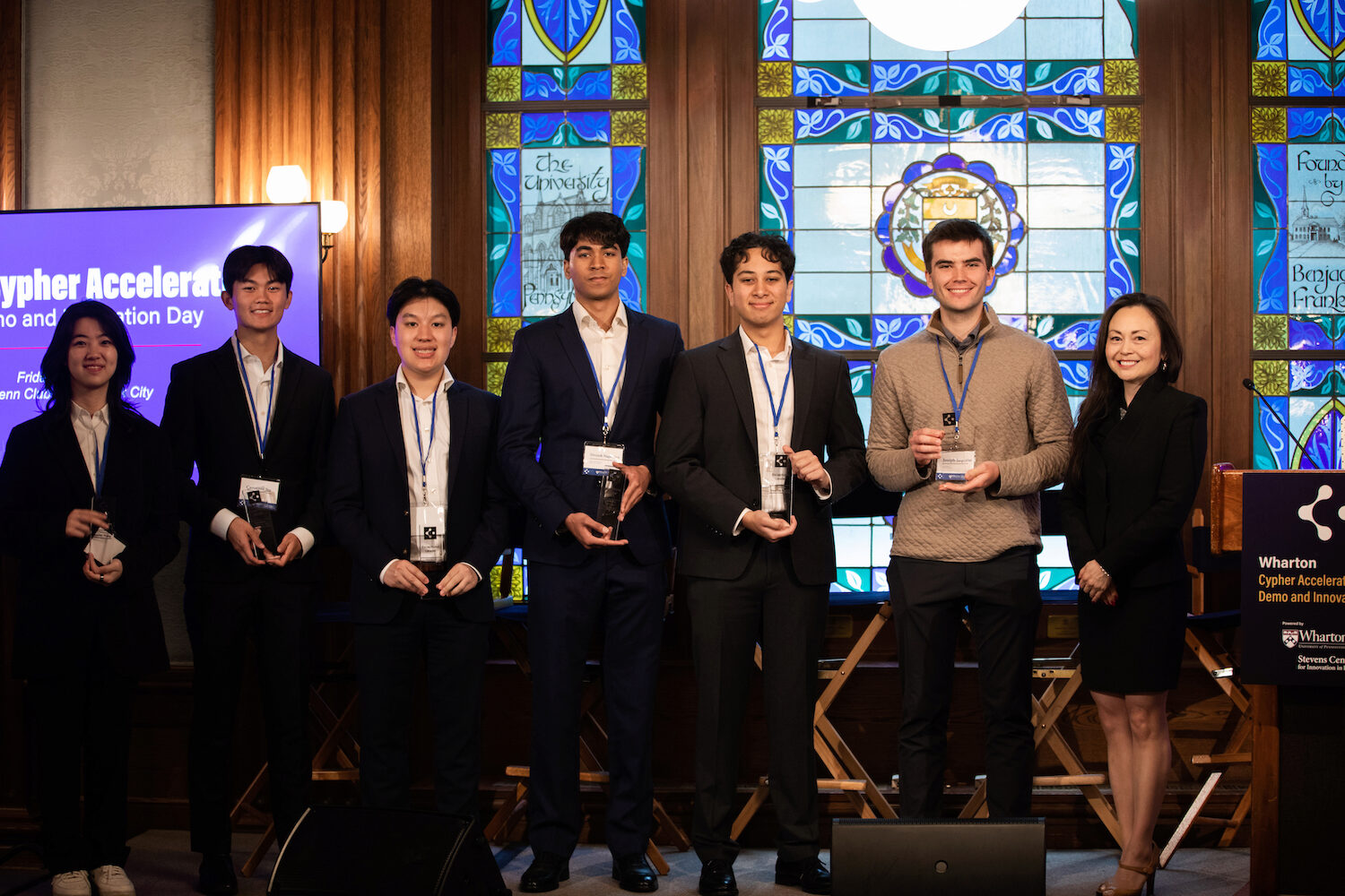 A group of people standing in front of a stained glass window, holding awards. They are dressed in formal attire, and a sign is visible with "Wharton Cypher Accelerator" text.
