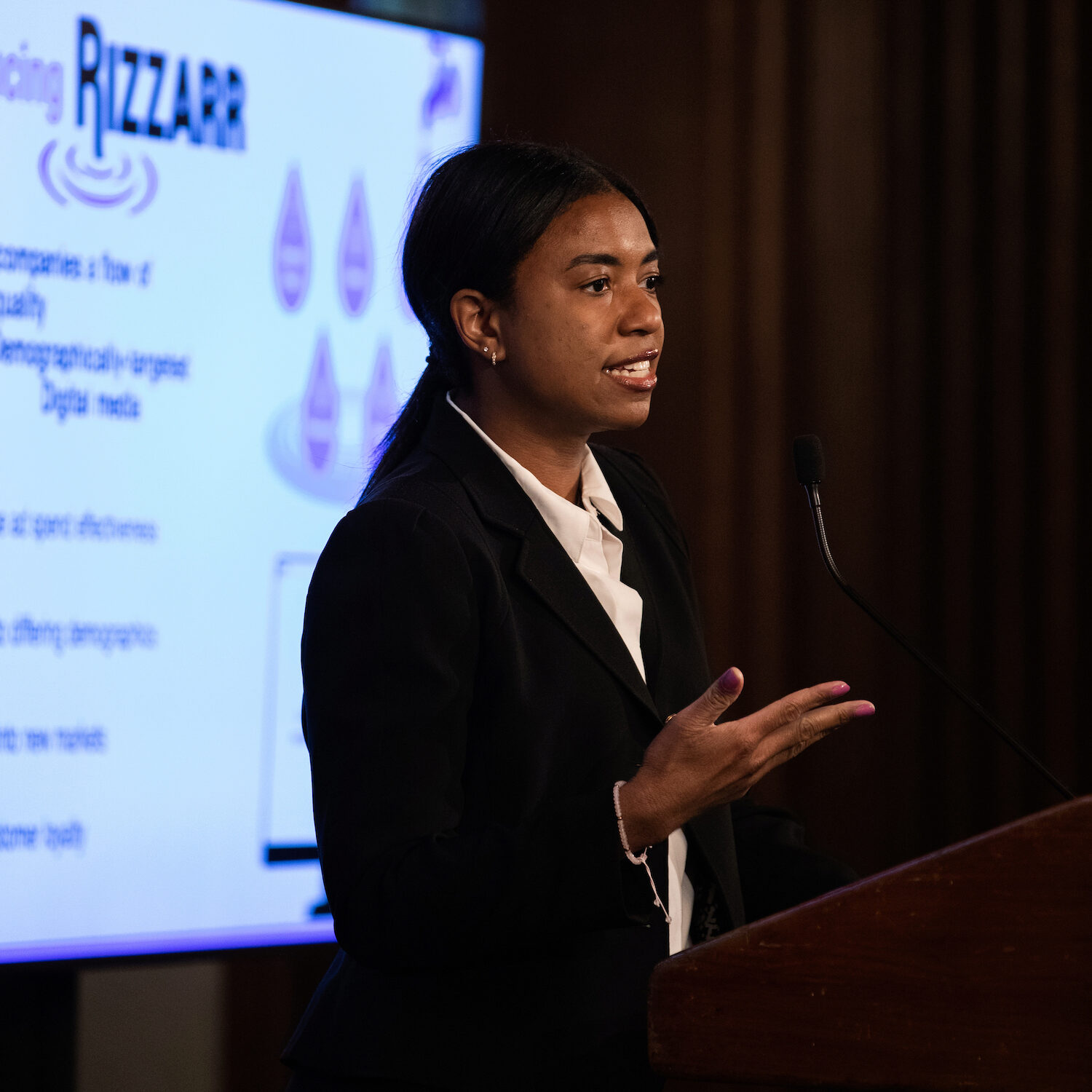 A speaker at a podium in front of a white screen that says "RIZZARR"