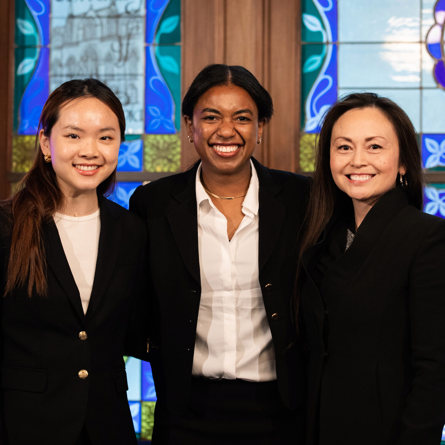 Three people pose for a group photo in front of stained glass windows, smiling toward the viewer