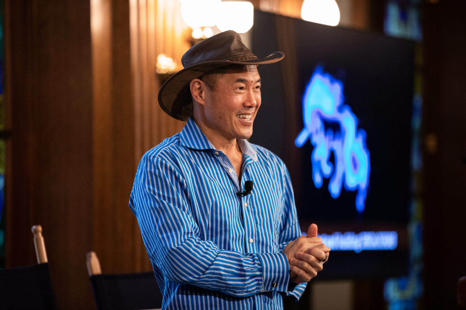 A speaker in a blue shirt and fedora-style hat smiles and holds his hands together while giving a keynote address.